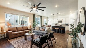 A spacious living room with a brown leather sofa, a gray sofa, and a coffee table. There is a ceiling fan, a large window with curtains, and a kitchen area in the background. - Park City Redbud Hideaway | Modern 6BR Mountain Retreat w/ Hot Tub & Views (Park City)