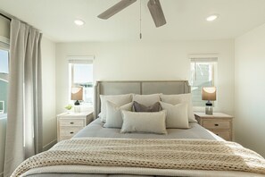 Une chambre à coucher comprenant un lit, deux tables de chevet, un ventilateur de plafond et des fenêtres équipées de stores.
