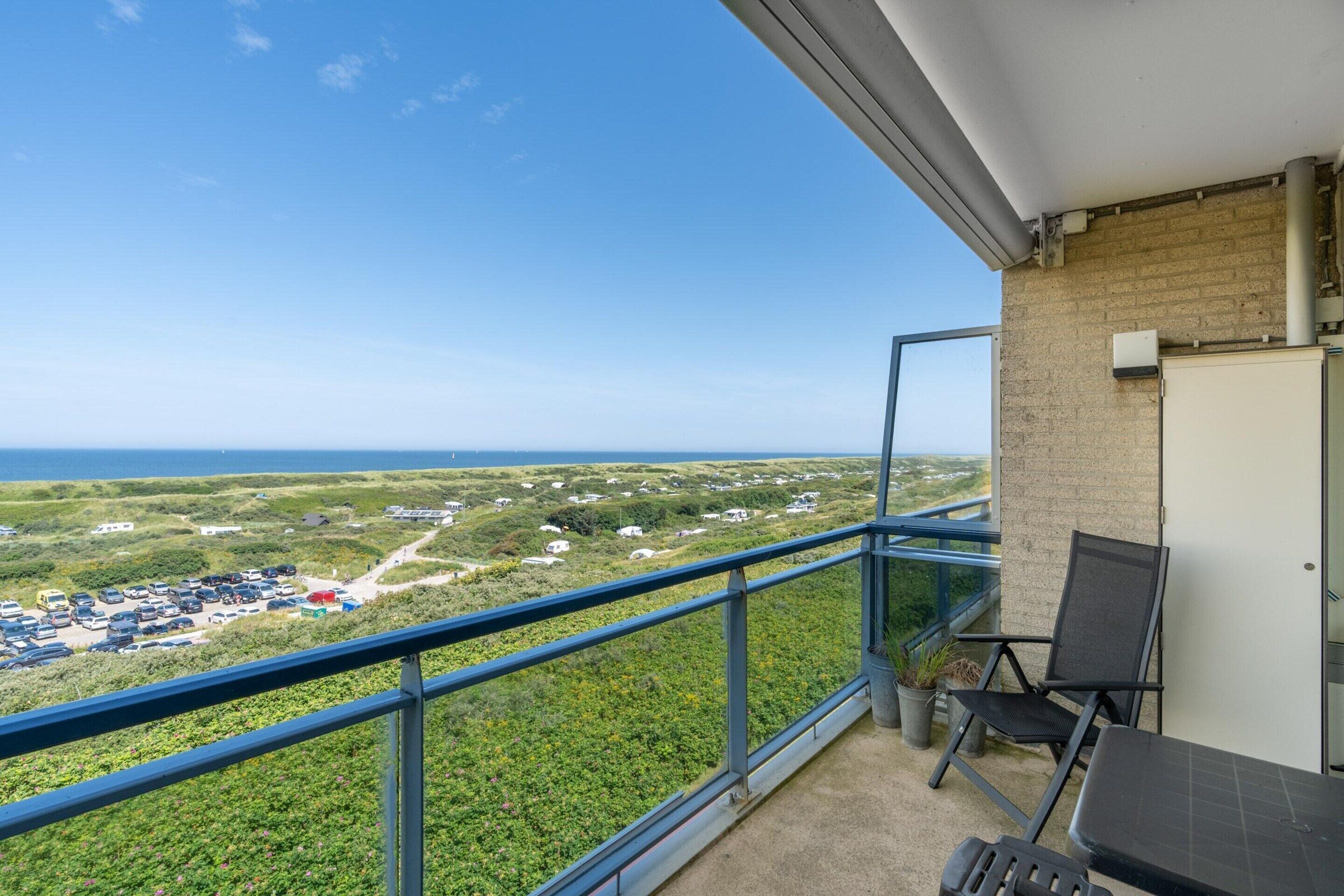A balcony with a view of a parking lot and greenery, a table, and a chair.