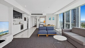 Un salon moderne avec un canapé, une table basse et un téléviseur. Il y a un coin cuisine avec des meubles blancs et un coin repas avec une table et des chaises.