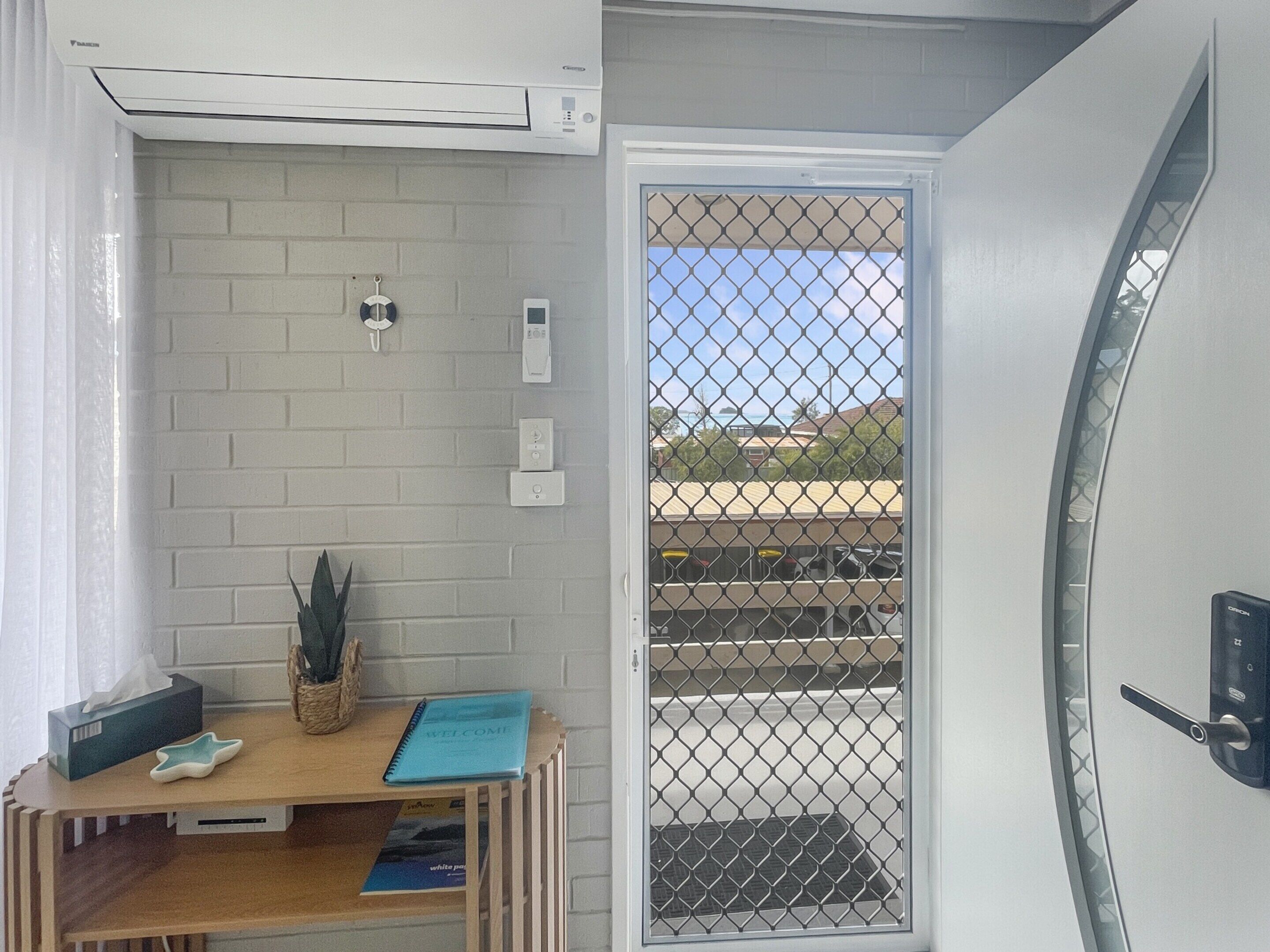 A room with a white brick wall, a wooden desk with a notebook, a tissue box, and a basket. There is a door with a metal screen, and an air conditioning unit above it.