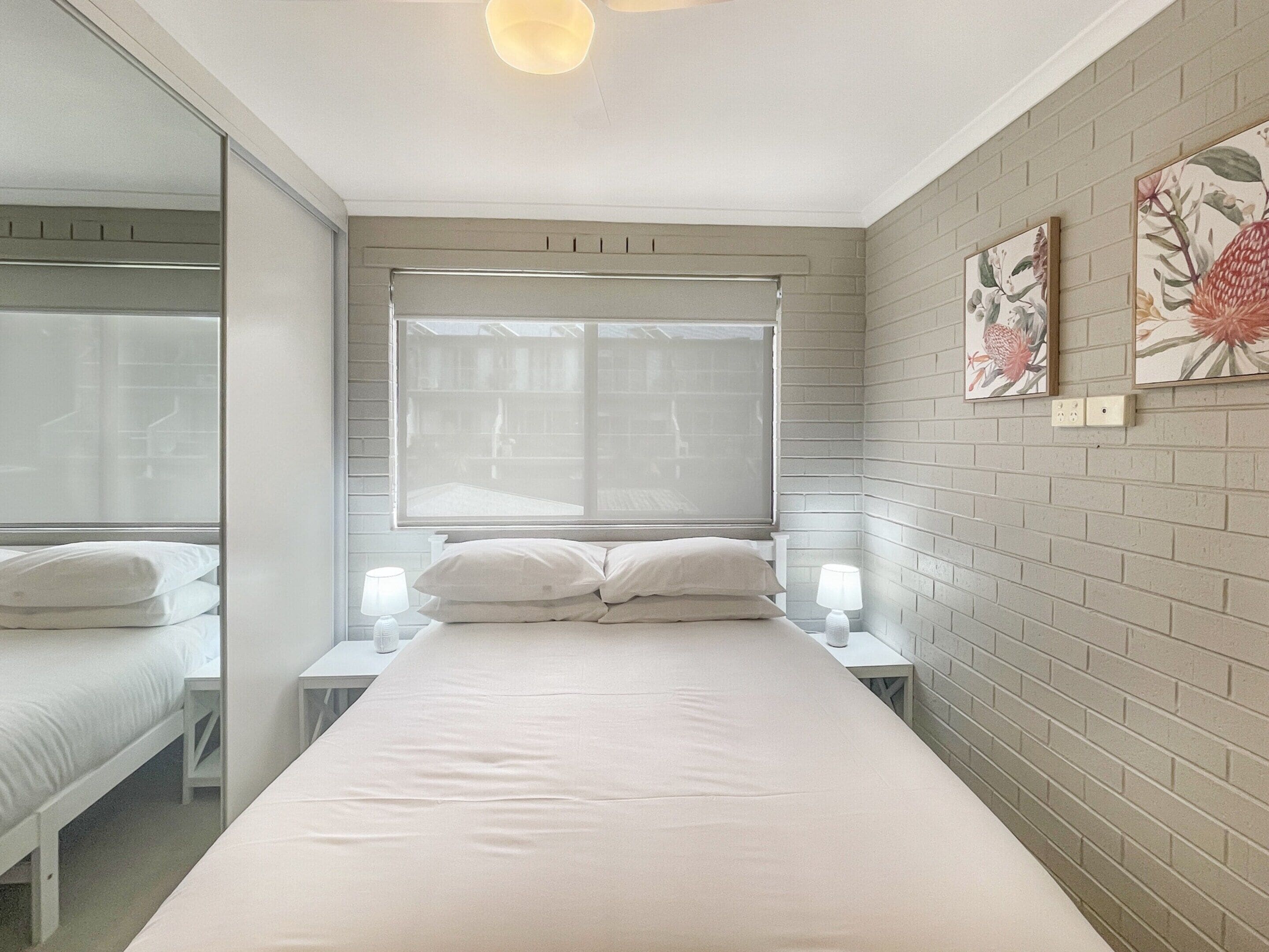 Een slaapkamer met een groot bed, twee nachtkastjes met lampen, een raam met jaloezieën en twee bloemschilderijen aan de muur.