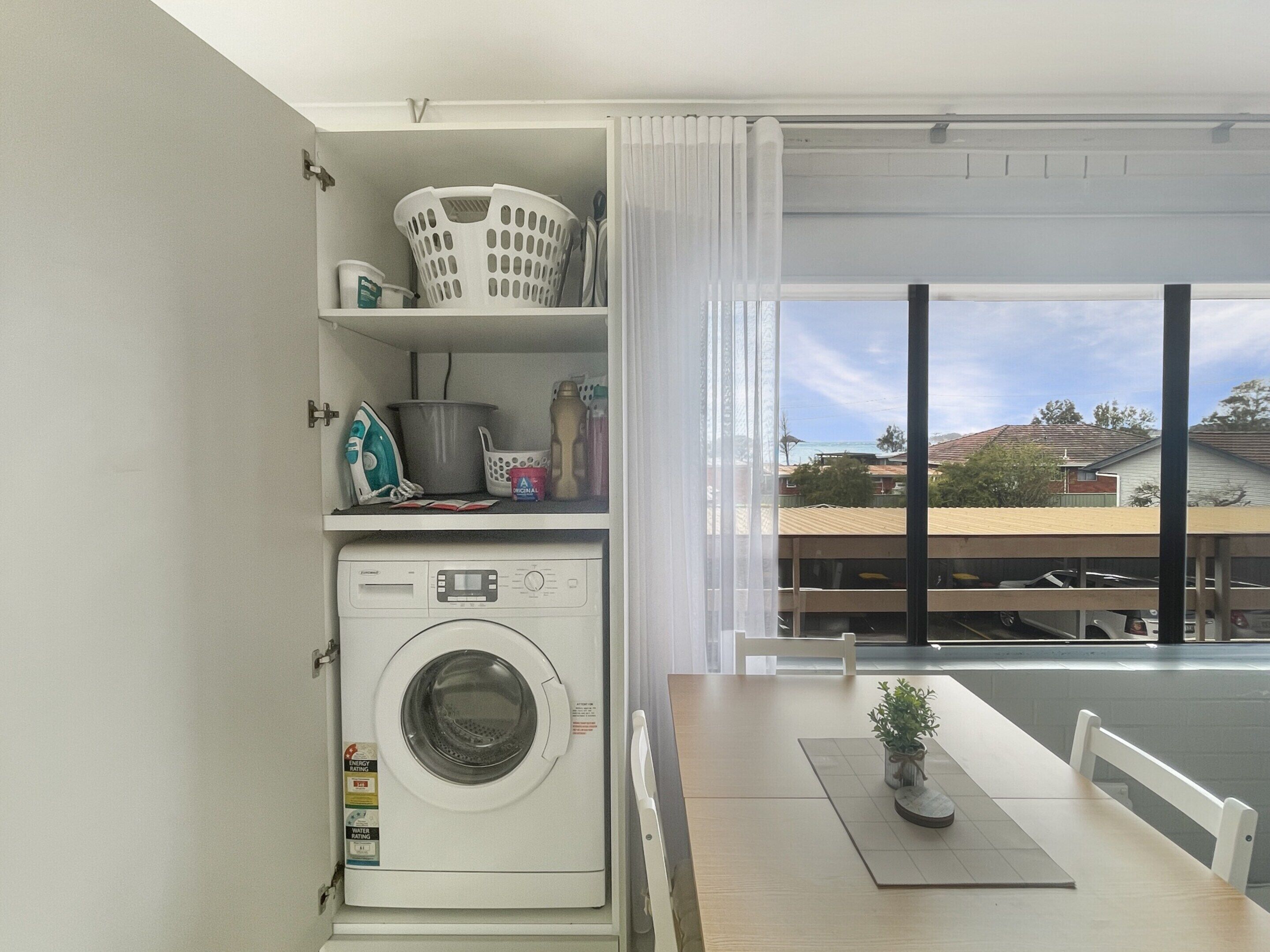 Een wasserette met een wasmachine, planken met wasmiddelen, een eettafel met een plant en een uitzicht op gebouwen door grote ramen.