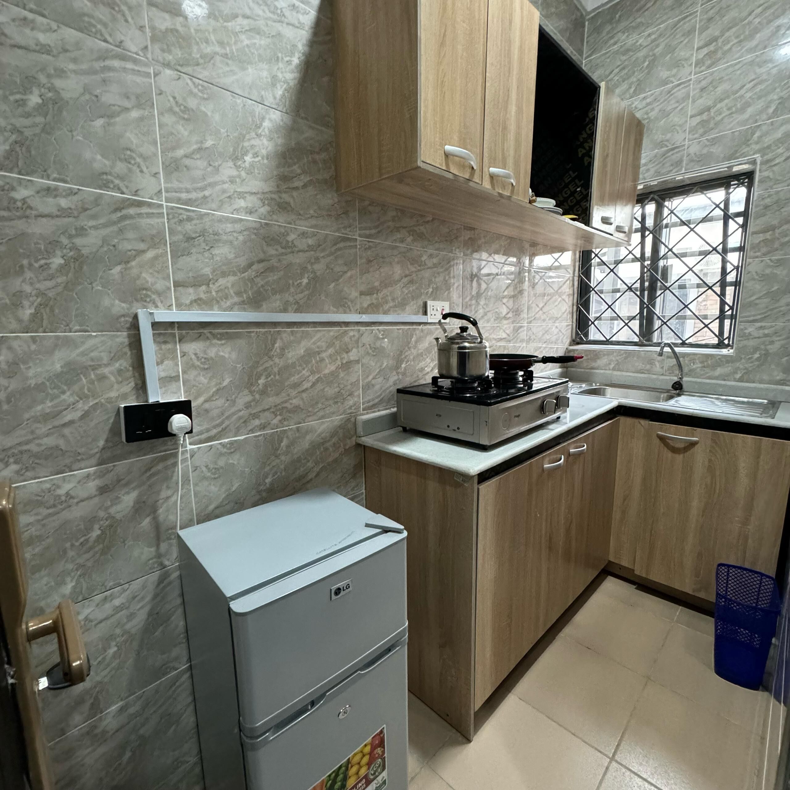 Eine kompakte Küche mit eingebautem Gaskochfeld, einem kleinen Kühlschrank, Holzschränken, einem Fenster mit Metallgitter und einer gefliesten Spritzschutzwand.