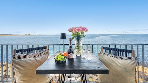 A table set with a bottle, a vase of flowers, and a bowl of fruit by a seaside view.