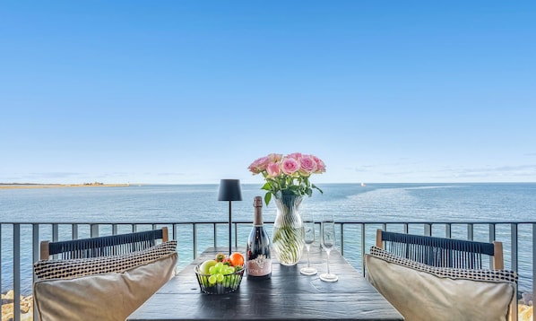 Uma mesa com uma garrafa, copos, um vaso de flores e uma cesta de frutas com vista para o mar.