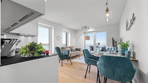 A modern kitchen with a dining area, a balcony with a view, and a living room with a sofa and chairs.