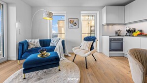 Un appartamento moderno con un divano blu, una poltrona bianca, una cucina con mobili bianchi e un'ampia finestra.