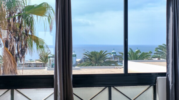 A view from a room with a large window overlooking a beach and palm trees. - Casa Ondina - Shared pool and Sea view By LVH (Puerto del Carmen)