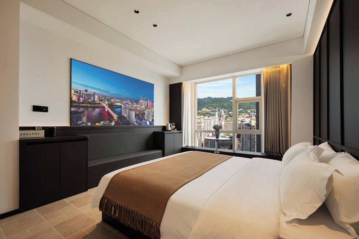 A hotel room with a large bed, a cityscape view through a window, a flat-screen TV on the wall, and a small table with a plant.