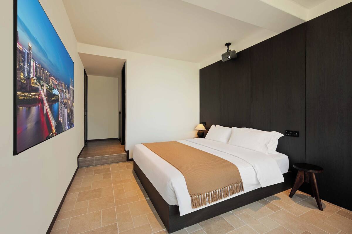 A modern hotel room with a large bed, a dark accent wall, a nightstand, and a wall-mounted projector.