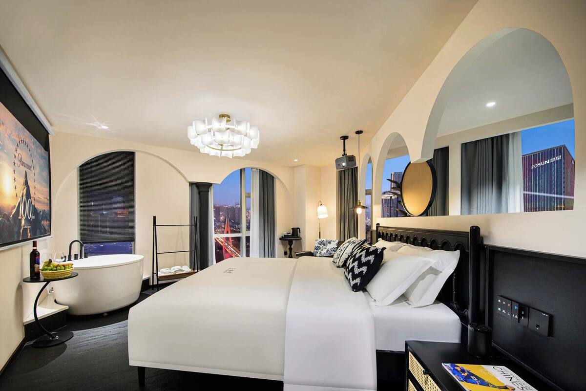 A modern hotel room with a large bed, a bathtub, and a city view.