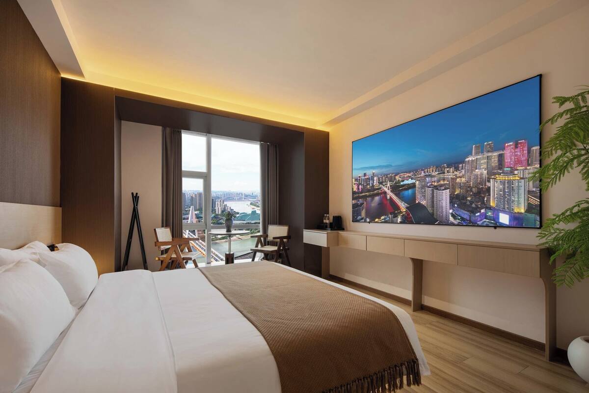 A hotel room with a large bed, a desk, a television, and a view of a cityscape.