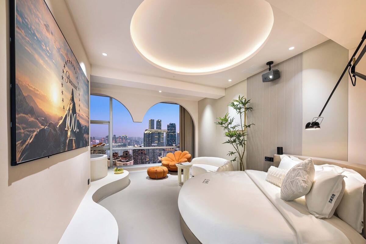 A modern hotel room with a large bed, a seating area with a view of the city, and a flat-screen TV mounted on the wall.