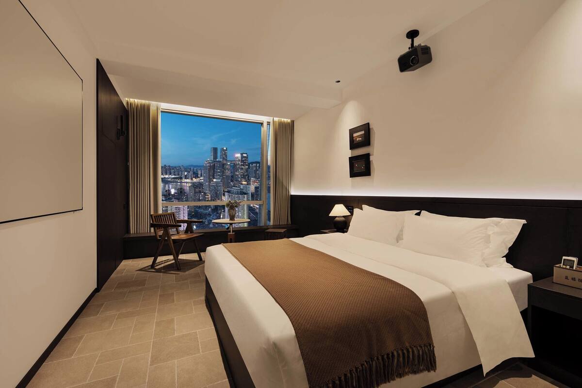 A hotel room with a large bed, a nightstand, a lamp, a chair, a small table, and a view of a cityscape through a large window.