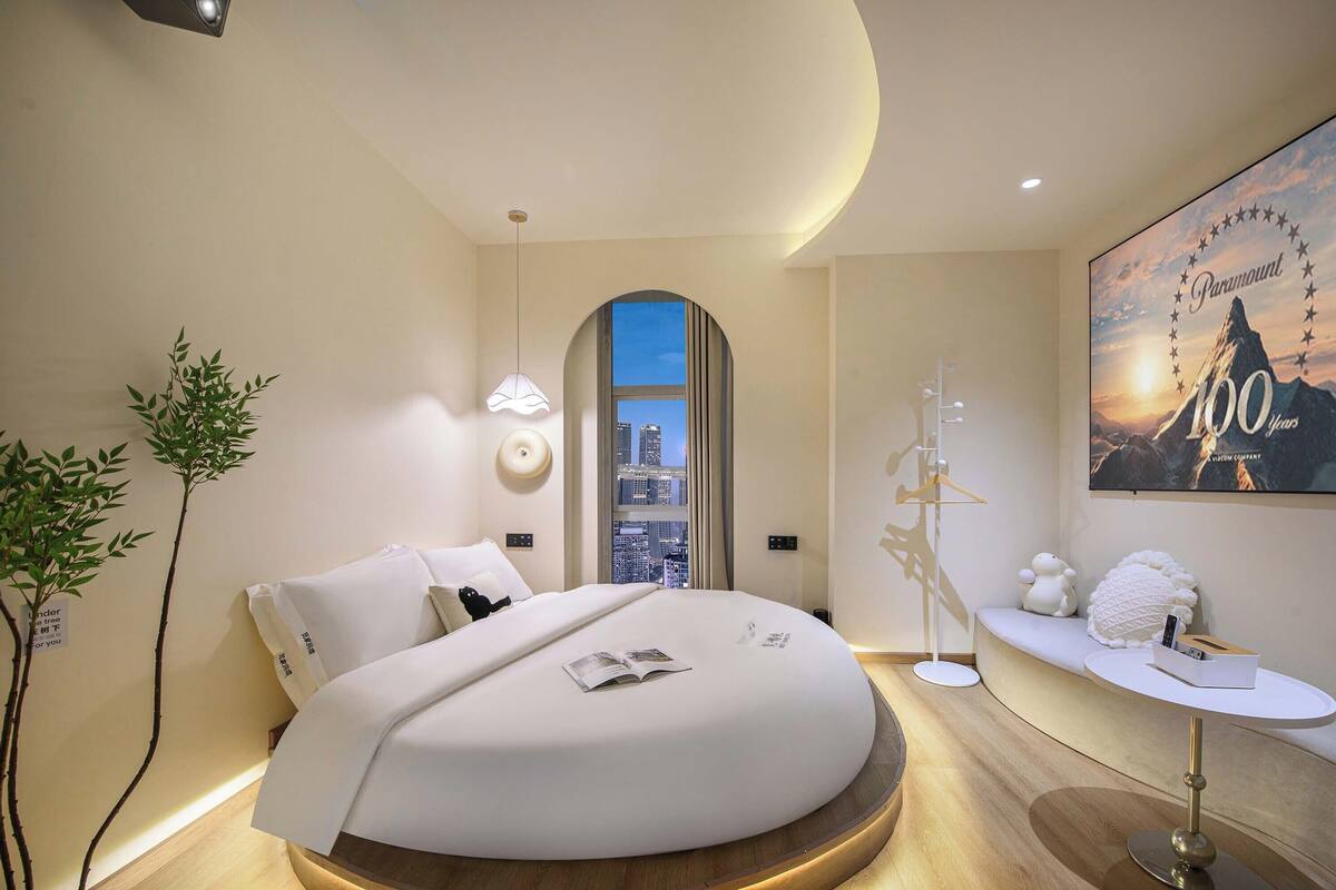 A modern hotel room with a curved bed, a city view through an arched window, a bedside table with a lamp, and a poster on the wall.