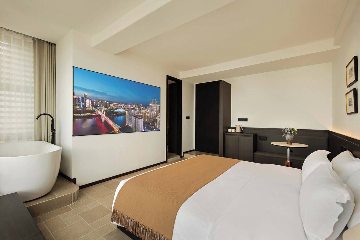 A modern hotel room with a large bed, a bathtub, a desk, and a cityscape view.