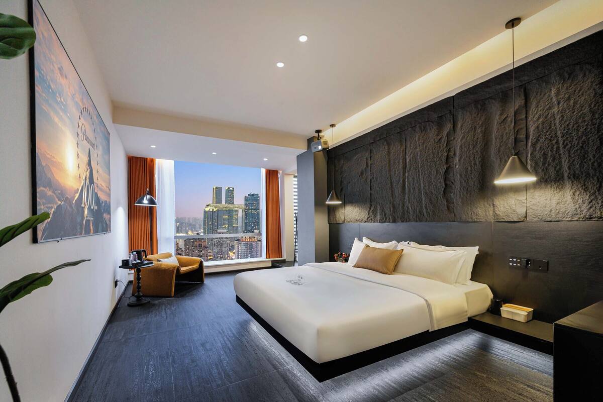 A modern hotel room with a large bed, a city view, a dark textured wall, and a small seating area with a lamp.