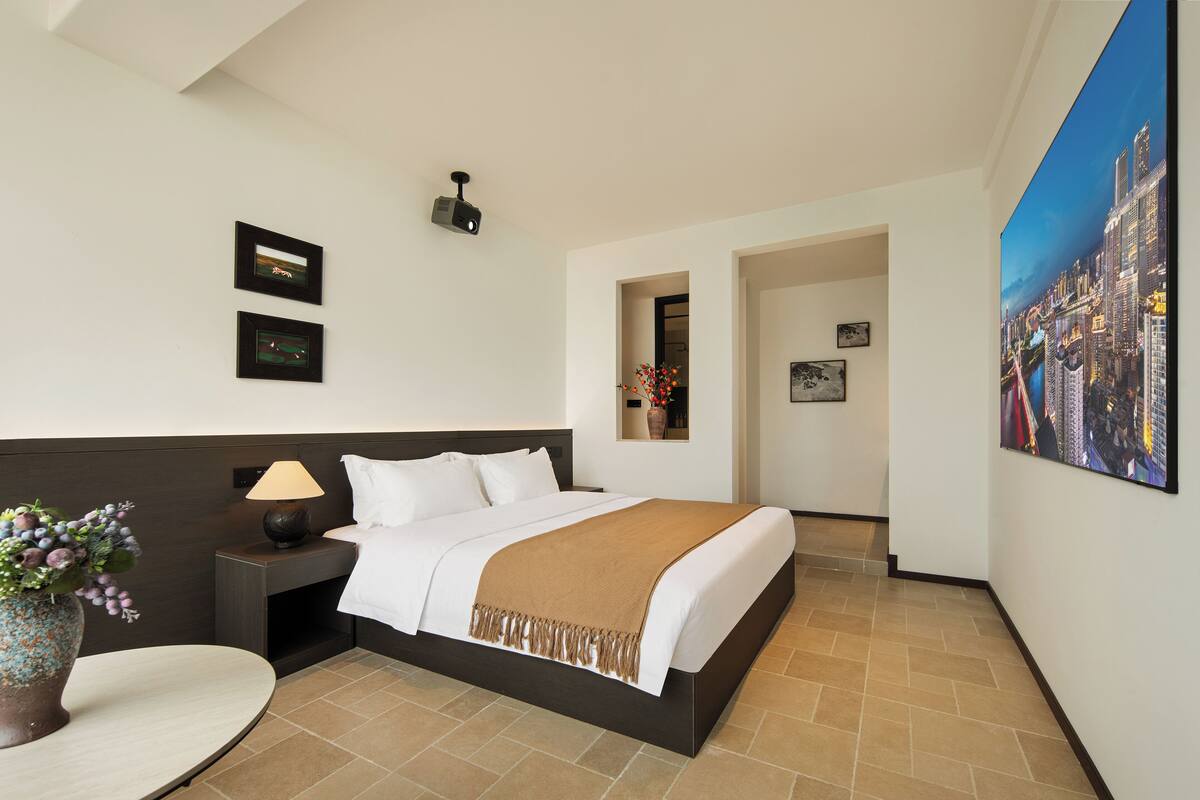 A hotel room with a bed, a nightstand, a lamp, a vase with flowers, and a painting of a cityscape.