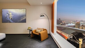 A modern hotel room with a large window offering a city view, a comfortable armchair, a small table with fruit, and a framed artwork on the wall. - Liberation Monument Riverside HoteL (Chongqing)