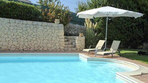 Ein Schwimmbecken mit Steinmauer, zwei Liegestühlen und einem weißen Sonnenschirm.