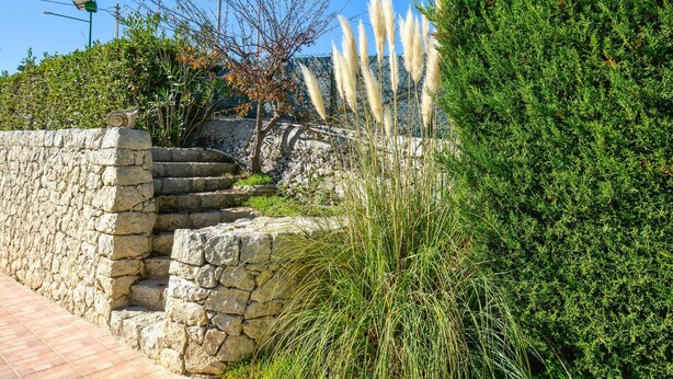 Eine Steintreppe führt zu einer Steinmauer, umgeben von üppiger Vegetation und Pampasgras.