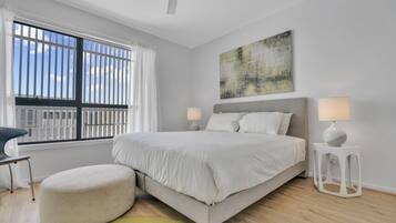 Un dormitorio moderno con una cama grande, dos lámparas de noche, un reposapiés redondo y vistas de la ciudad a través de la ventana.