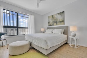 A modern bedroom with a large bed, two bedside lamps, a round ottoman, and a city view through the window. - Oceanview Luxury With Dedicated Workspace (Adelaide)