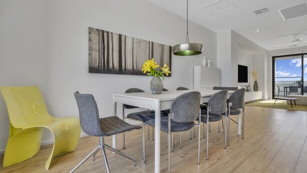 A modern dining area with a white table, grey chairs, and a yellow chair. A painting of a forest is on the wall, and a vase with yellow flowers is on the table. - Oceanview Luxury With Dedicated Workspace (Adelaide)