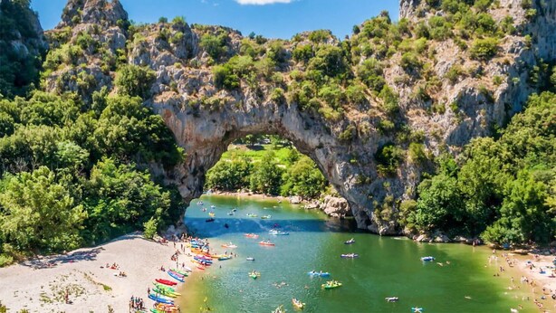Une arche naturelle formée par la roche, enjambant une rivière où l’on voit des bateaux et des personnes sur la rive.