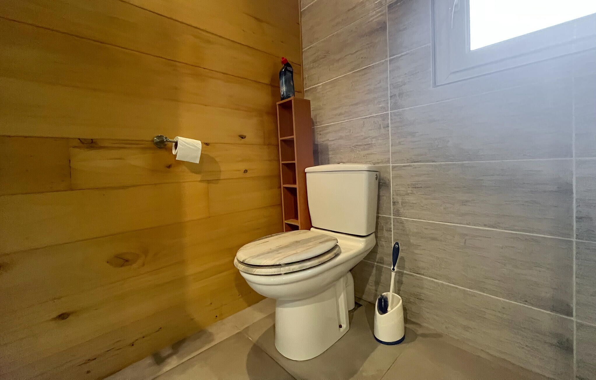 Une salle de bain aux murs lambrés, avec des toilettes, une étagère sur laquelle se trouve une bouteille et une brosse à toilettes.