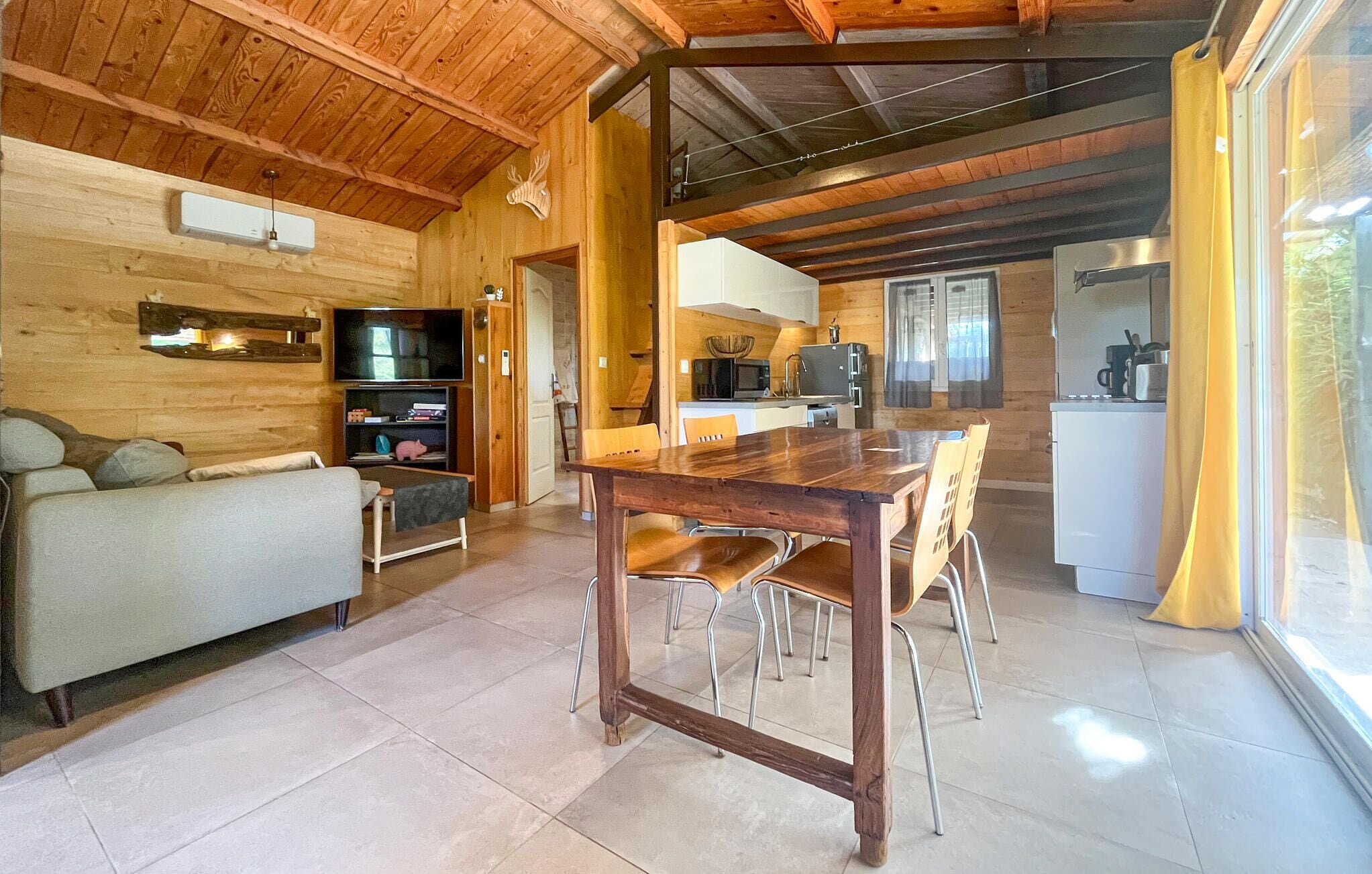 A cozy log cabin interior with a wooden dining table, chairs, a sofa, and a kitchen area with a refrigerator and microwave.