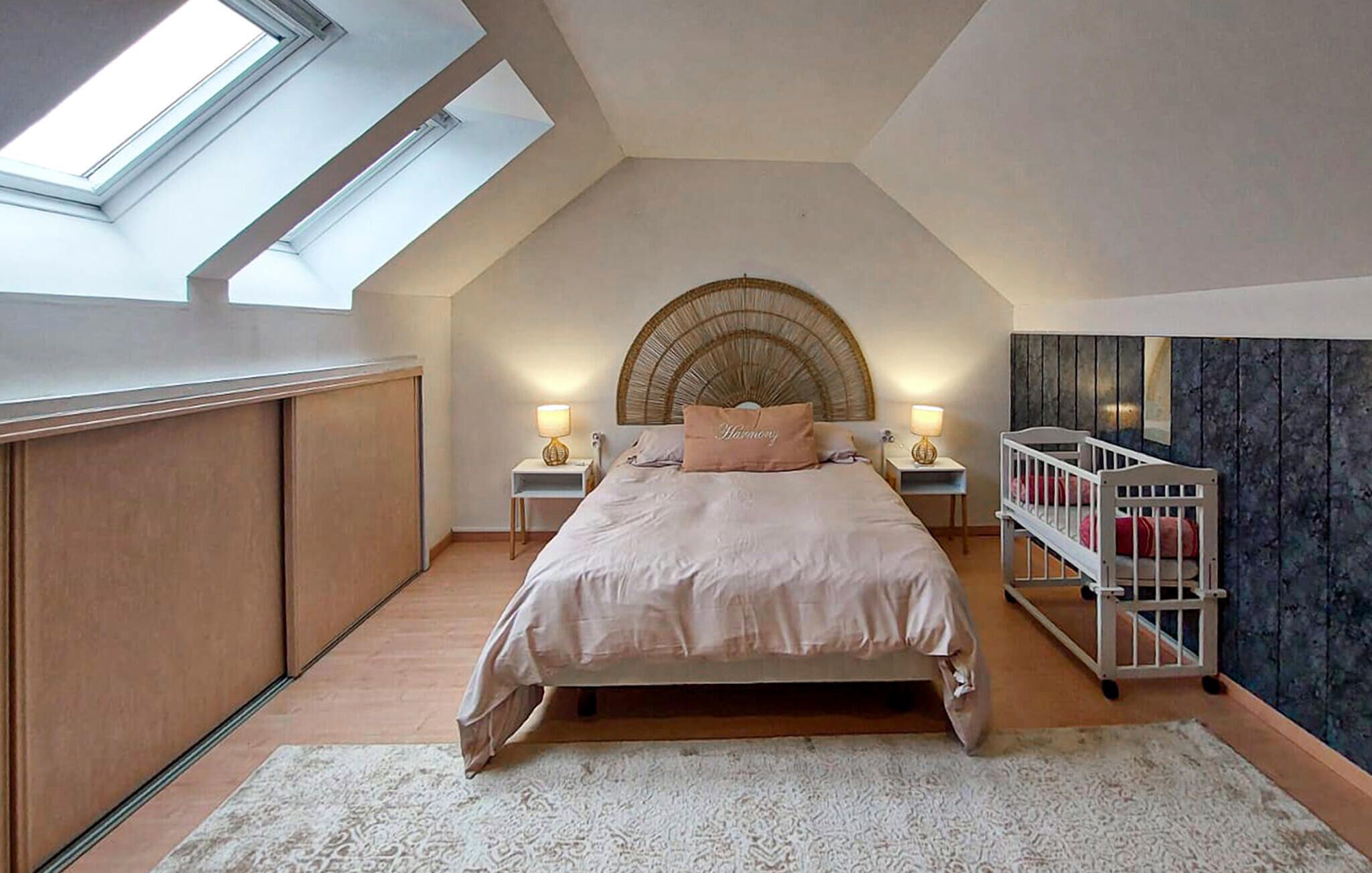 A bedroom with a skylight, a bed with a headboard, bedside tables, a crib, and a textured wall.