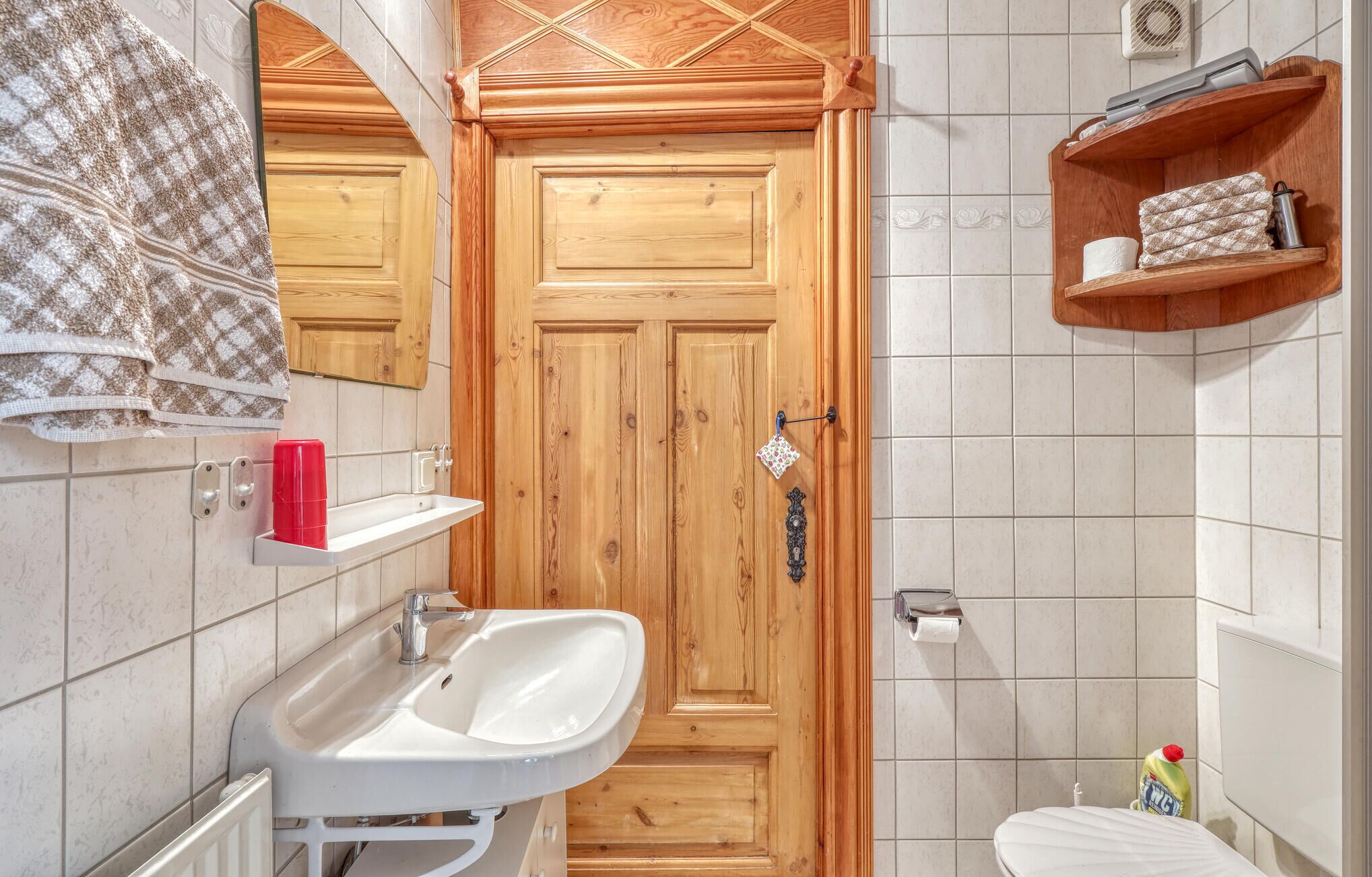 Een badkamer met een houten deur, een witte wasbak, een toilet en een plank met toiletartikelen.