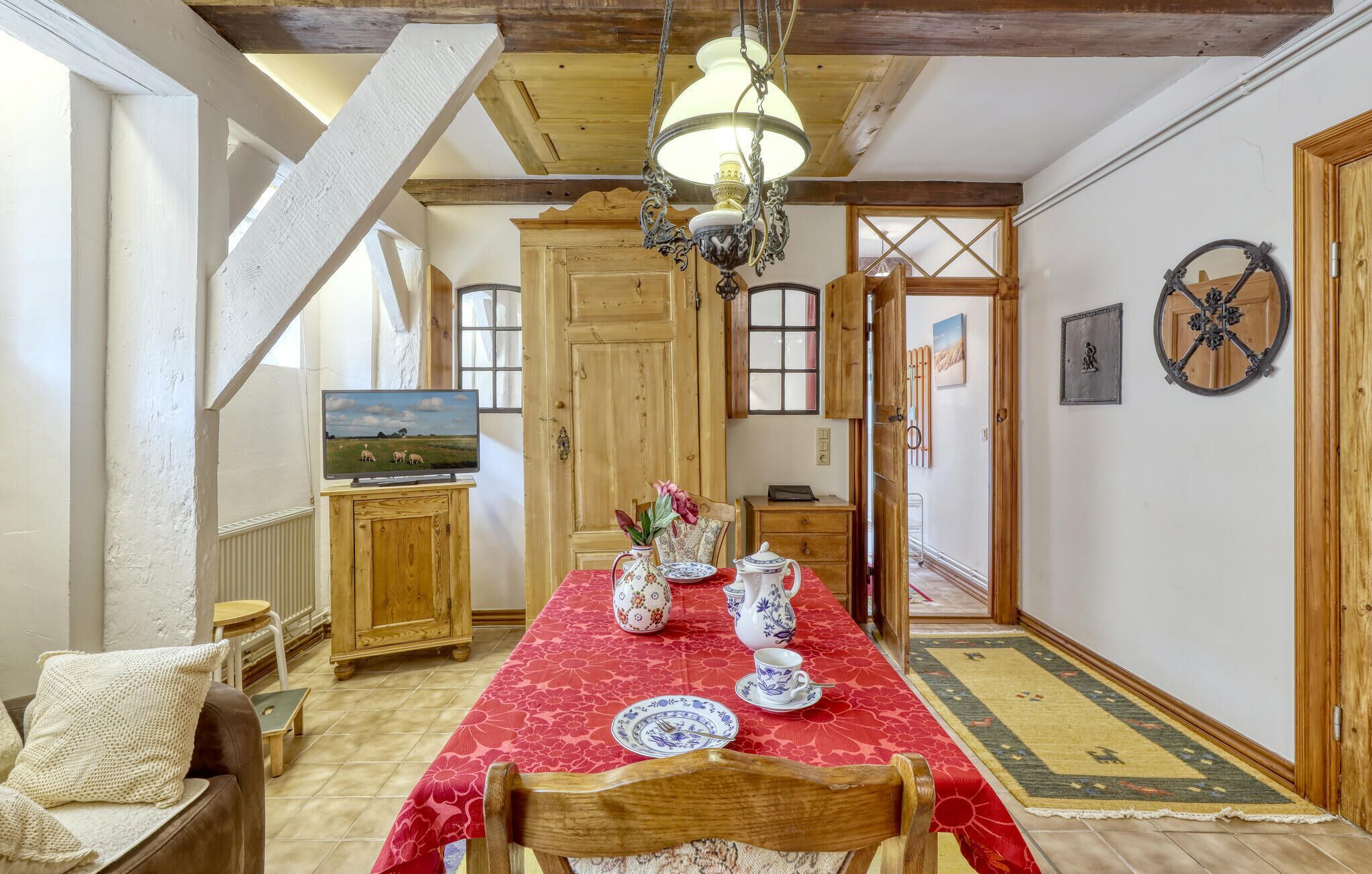 A dining area with a red tablecloth, wooden chairs, and a chandelier. A television is mounted on a cabinet, and there are wooden doors and a rug in the background.