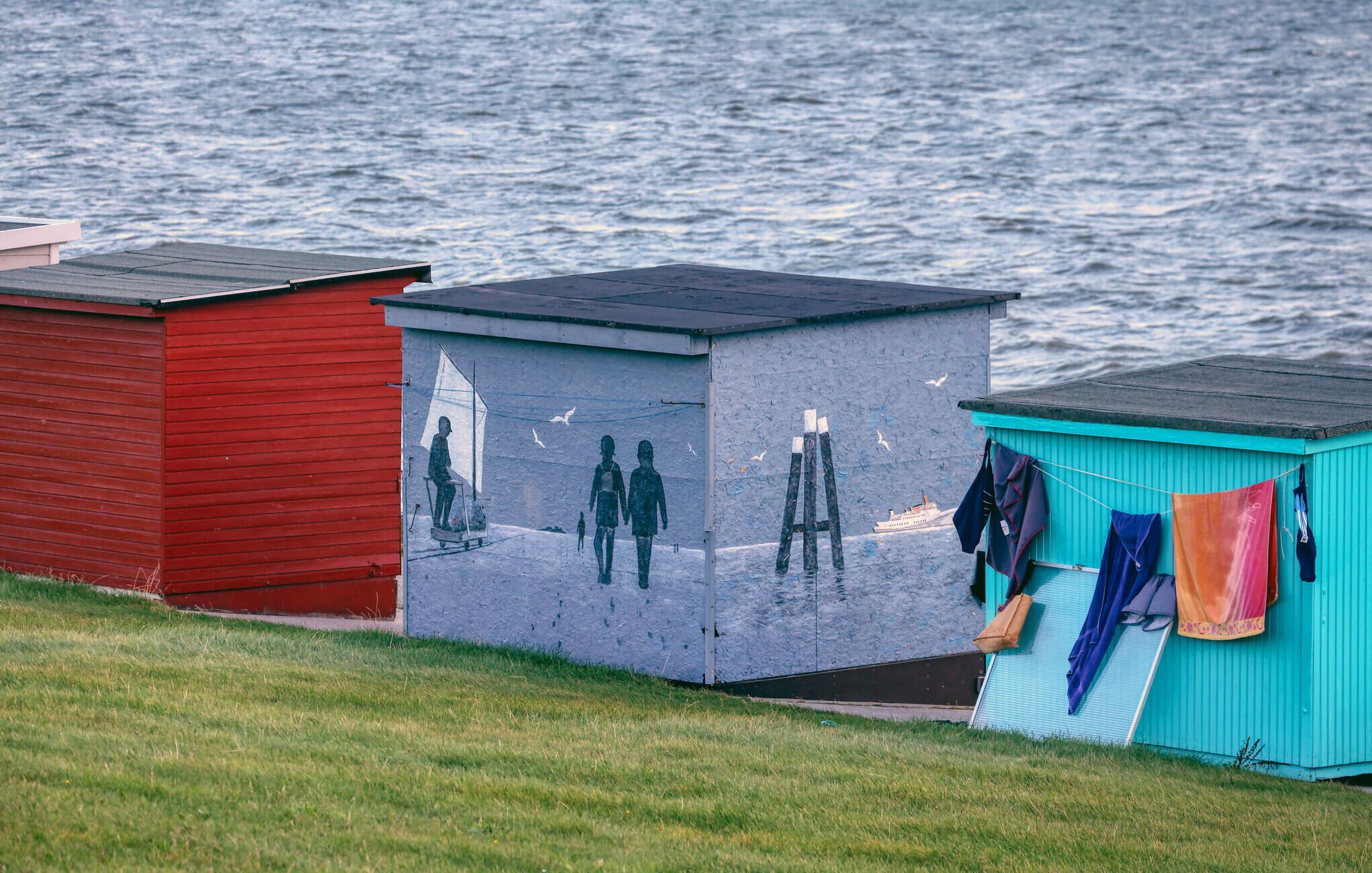 Eine Reihe kleiner, bunter Schuppen am Wasser, wobei einer davon eine Wandmalerei mit Menschen und einem Segelboot zeigt.
