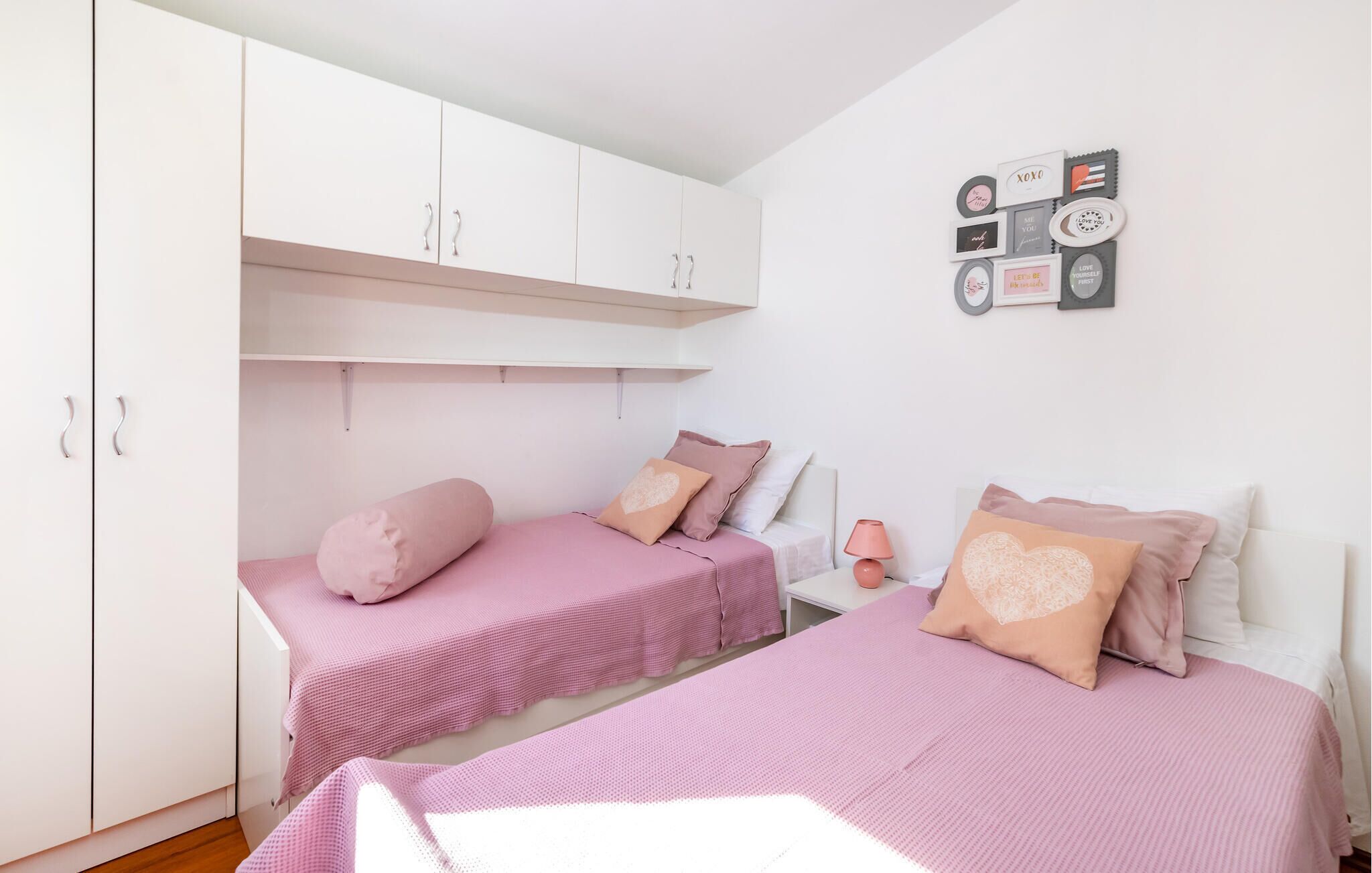 Un dormitorio con dos camas, cada una con ropa de cama rosa y almohadas con forma de corazón. Hay un armario blanco con asas plateadas y una mesita de noche con una lámpara.
