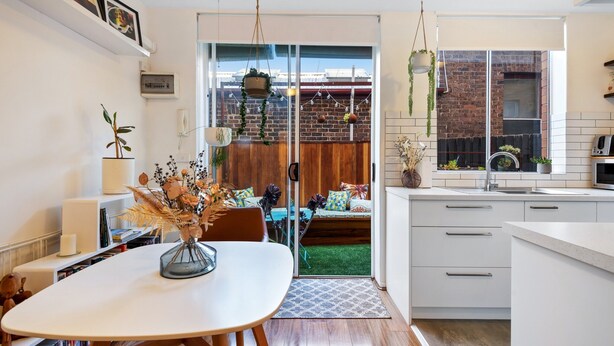 A modern kitchen with white cabinetry, a dining area with a white table and chairs, and a view of a brick wall and greenery outside. - Snug St Kilda Apartment With Private Courtyard (Melbourne)