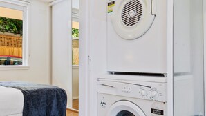 A laundry room with a stacked washer and dryer, a bed with a blue blanket, and a view of the outside through a window. - Snug St Kilda Apartment With Private Courtyard (Melbourne)