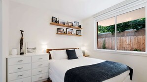 A bedroom with a bed, white nightstands, a dresser, and wall-mounted shelves with pictures. - Snug St Kilda Apartment With Private Courtyard (Melbourne)