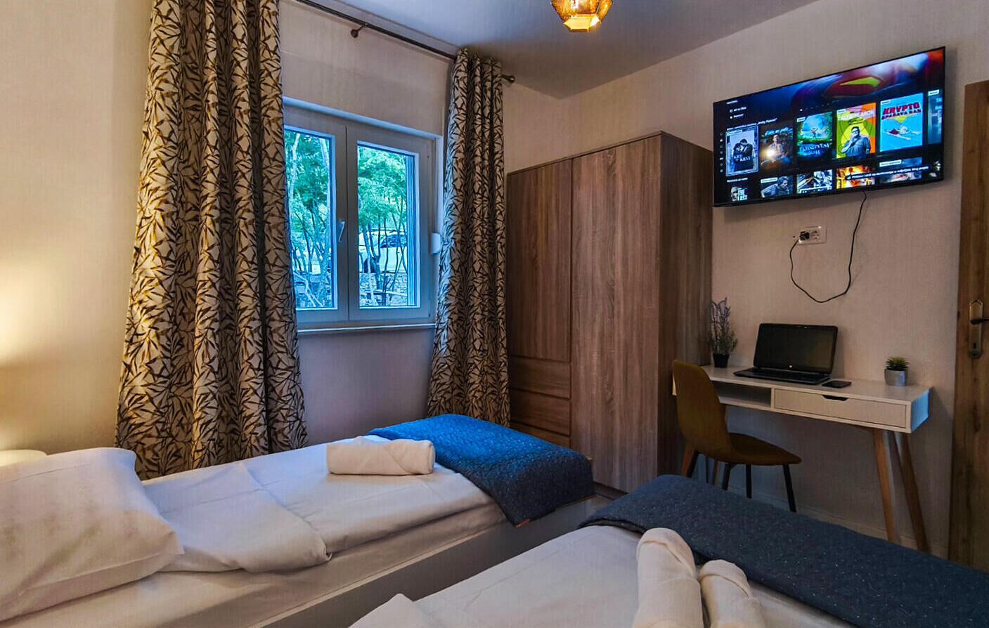 A room with two beds, a desk with a laptop, and a TV displaying a movie.