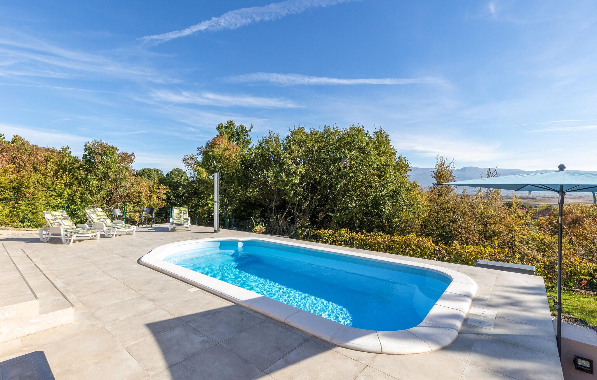 Piscina rectangular con solárium empedrado, rodeada de árboles y un cielo despejado.