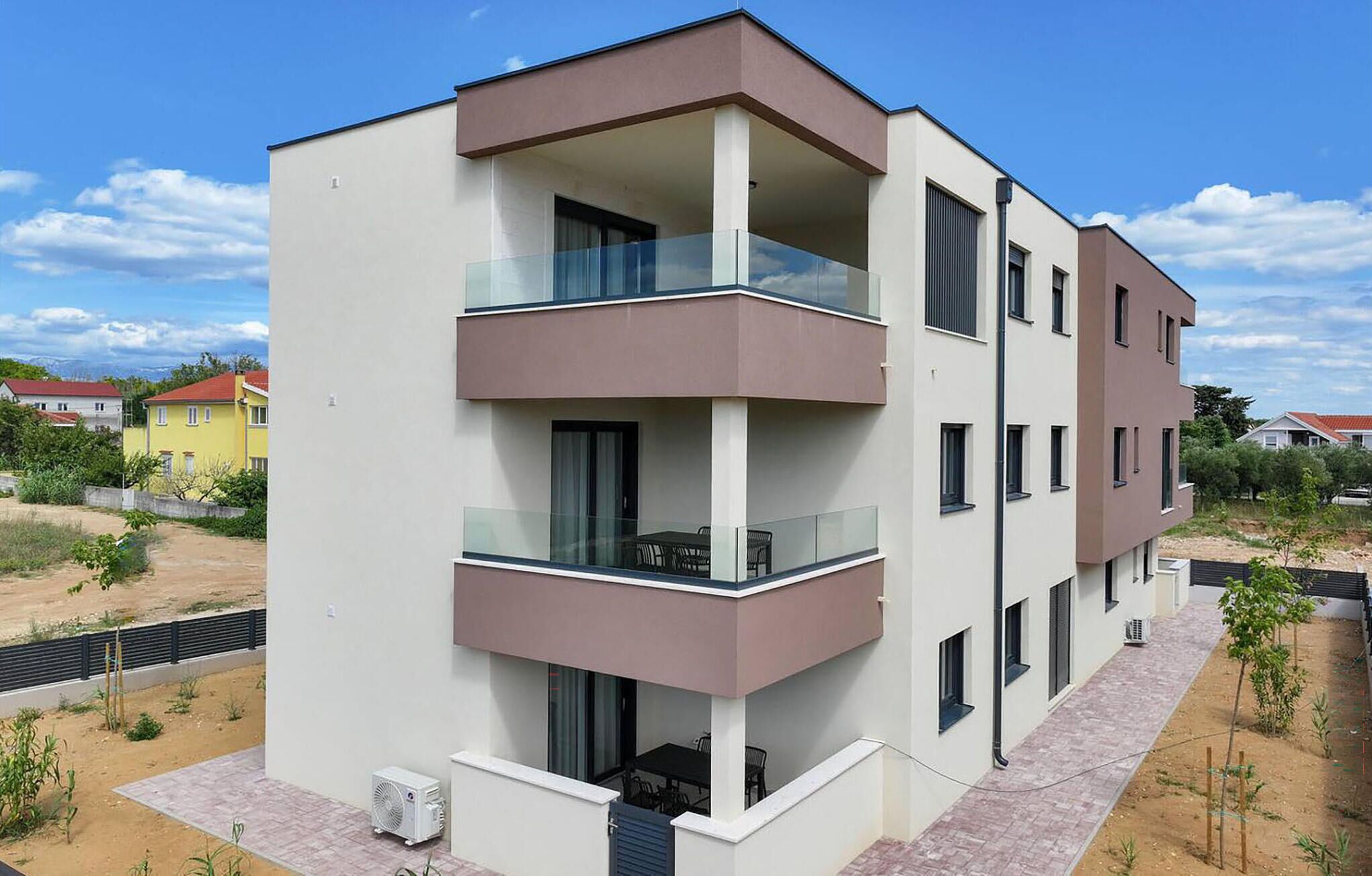 A modern multi-story residential building with balconies, a flat roof, and a paved ground area.