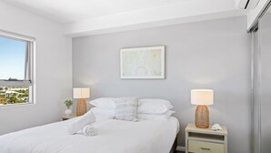 A modern bedroom with a large bed, two bedside tables with lamps, a wall-mounted artwork, and a window with a city view. - Seaside Escape With Pool, Balcony and Views (Redcliffe)