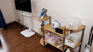 A hotel room with a flat-screen TV mounted on the wall, a wooden side table with a kettle, a digital clock, and a small cabinet with various items.