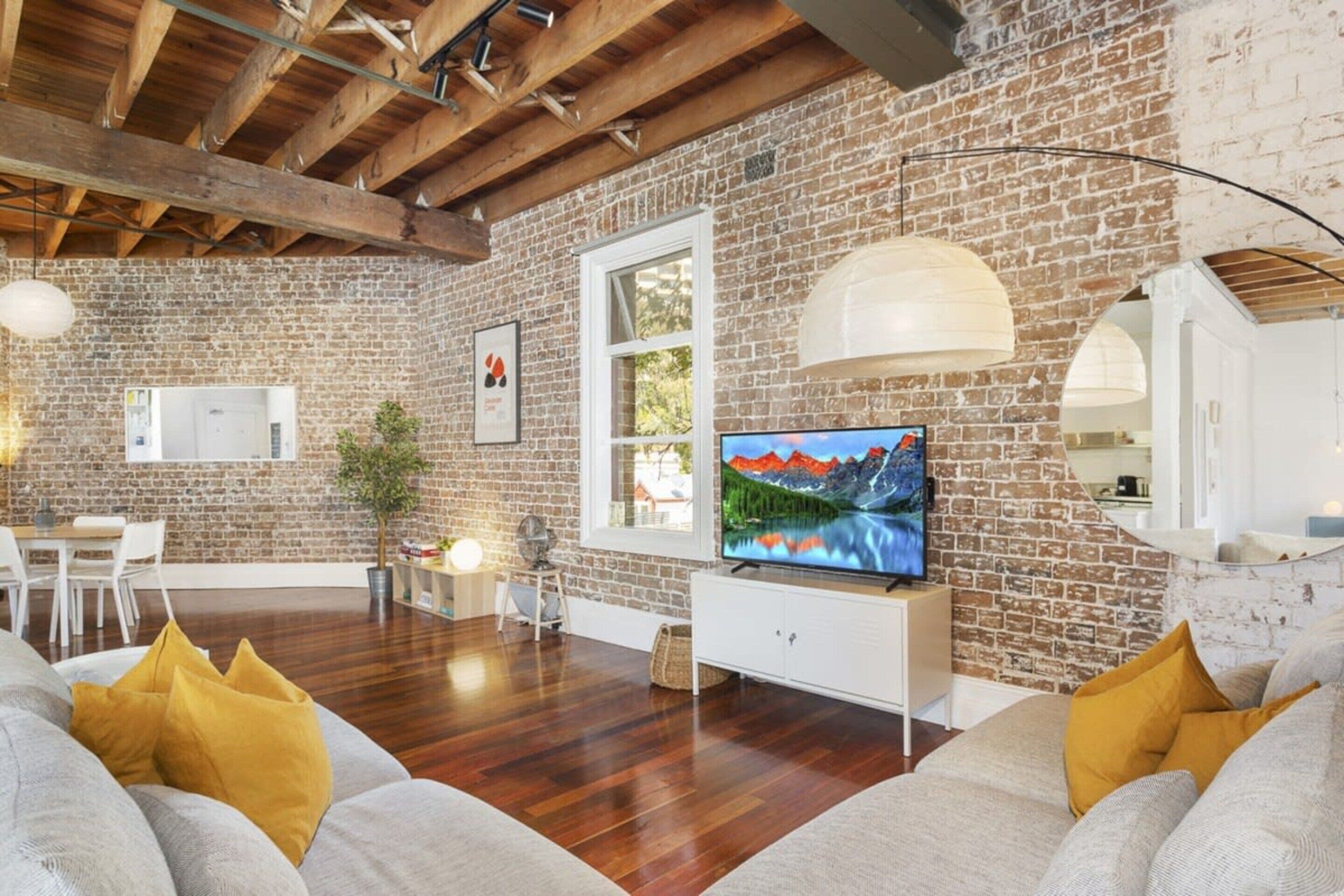 A spacious living room with a brick wall, wooden beams, a large flat-screen TV, and a dining area with white furniture.