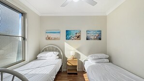 Une chambre avec deux lits simples, un ventilateur de plafond, une fenêtre donnant sur un immeuble et trois tableaux accrochés au mur.