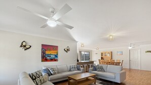 Un salon spacieux avec deux canapés, une table basse et un ventilateur de plafond. Au fond, on aperçoit un coin cuisine avec une table à manger et des chaises.
