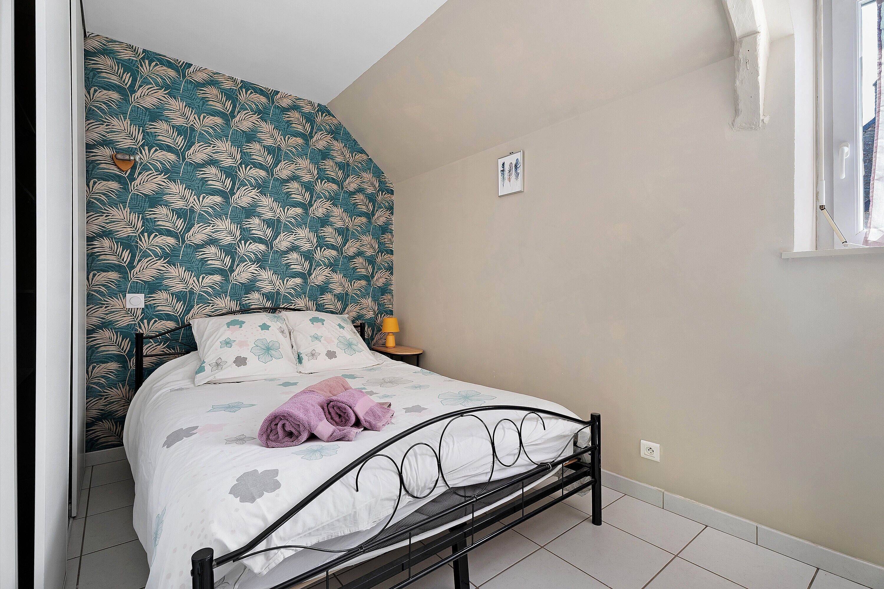 Un dormitorio con una cama, una mesita de noche, una ventana y un papel tapiz con diseño de hojas.