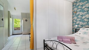 A bedroom with a bed, a window with curtains, a door, and a view of a green outdoor area. - Studio – Le Clos des Pommiers (Pleudihen-sur-Rance)